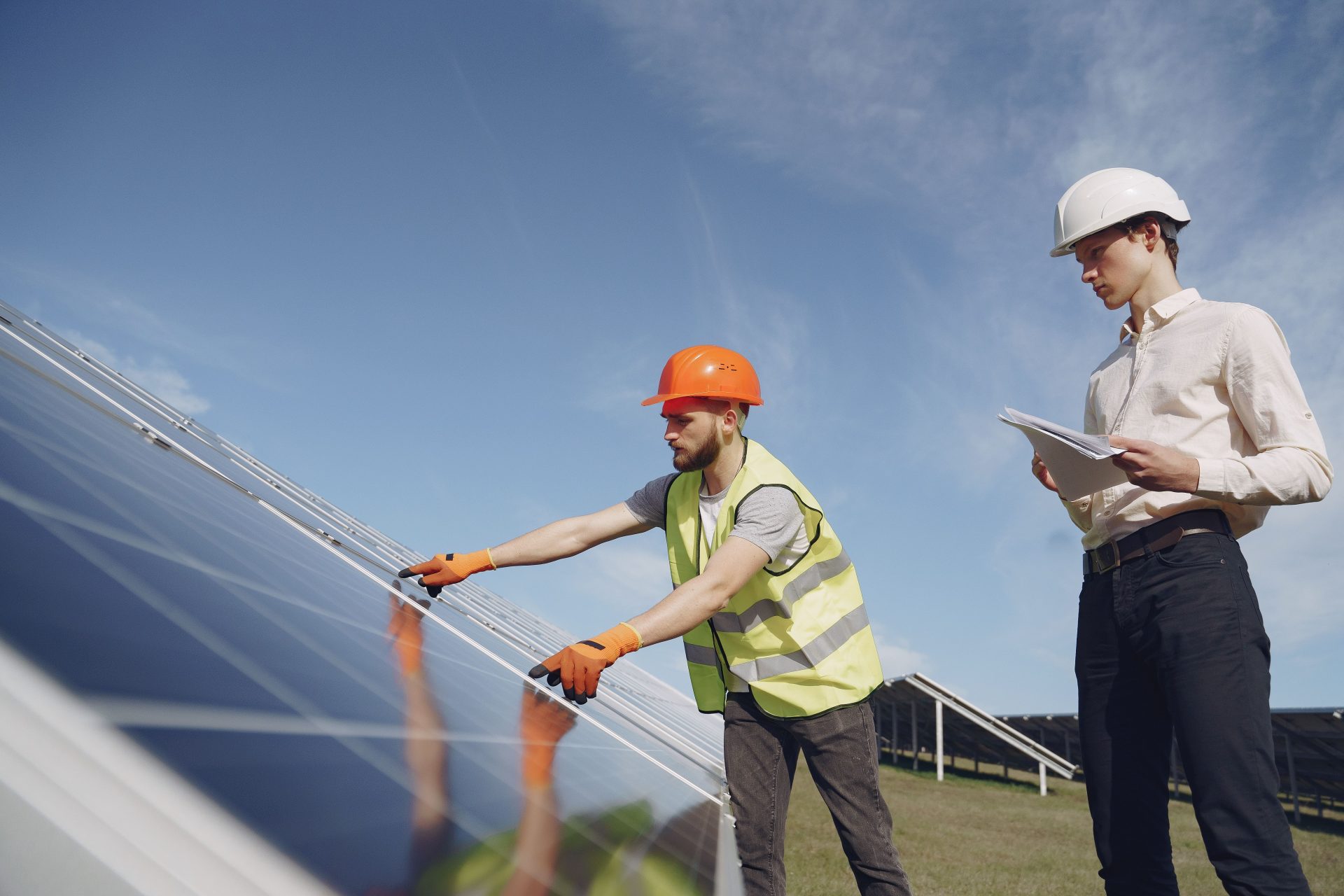 Ansicht von unten auf einen bärtigen Elektriker in gelber Weste und Schutzhelm, der einem jungen Inspektor in formeller Kleidung Solarmodule vorführt.